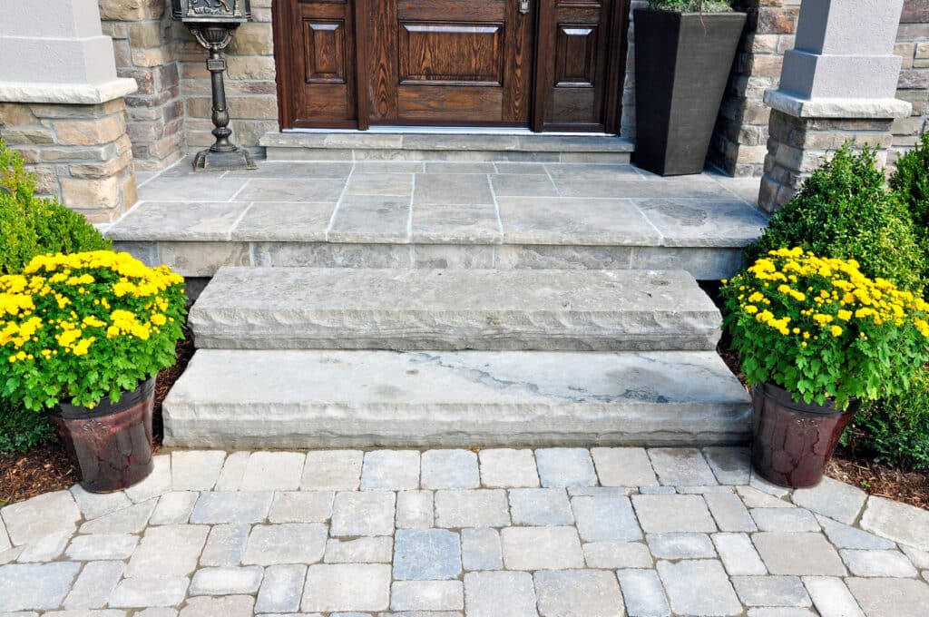 Flagstone applied to the original concrete veranda, natural stone steps, and paver walkway all provide a beautiful, fresh landscape update to this stately home.